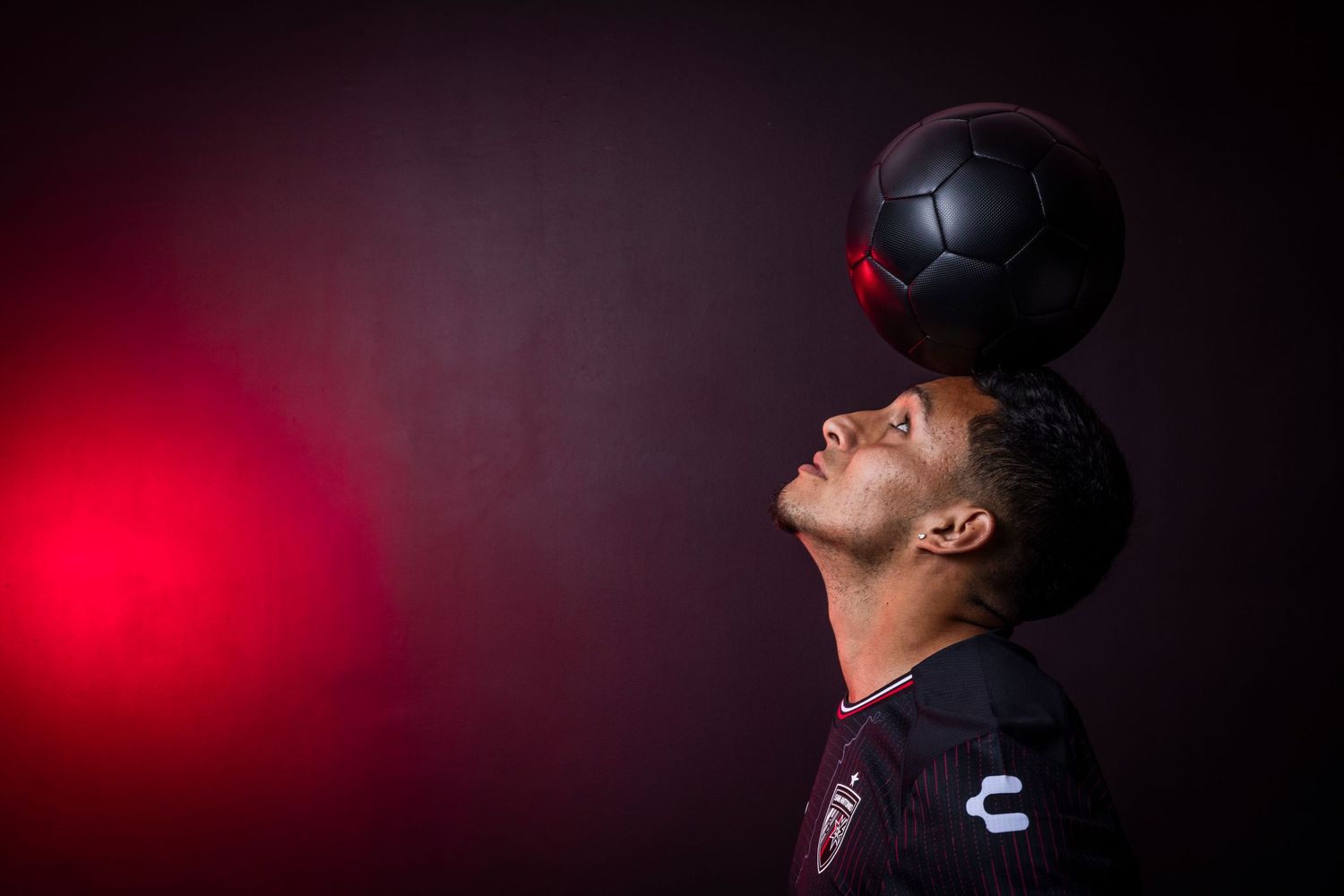 Soccer player balancing black ball on forehead against dramatic red lighting.