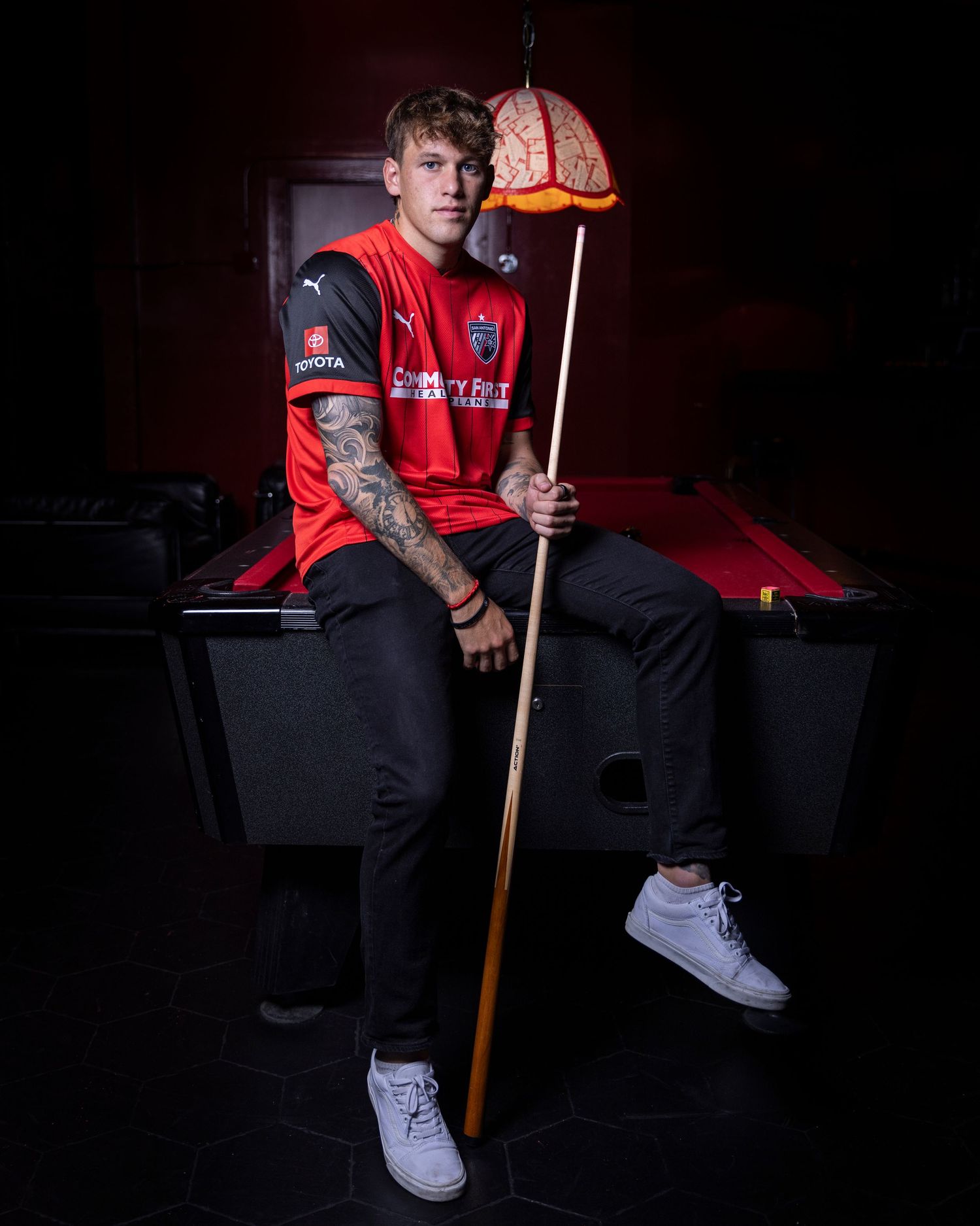 Athlete in red jersey sits on pool table holding cue stick under vintage pendant lamp.