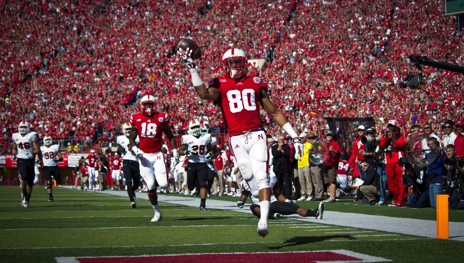 Football player celebrates touchdown by leaping with arms raised in stadium filled with red-clad fans.