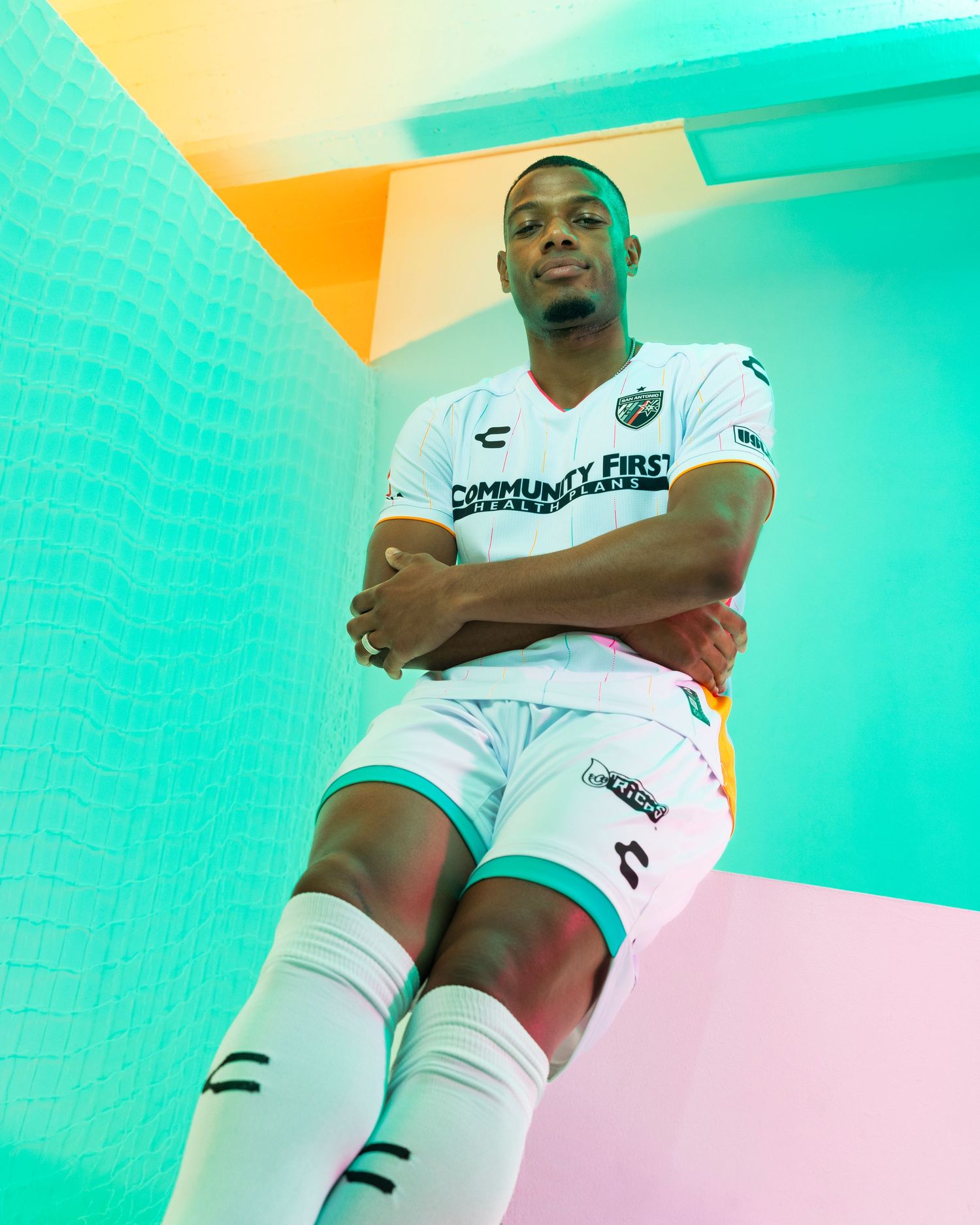 Soccer player poses in a white uniform against vibrant turquoise and orange walls.
