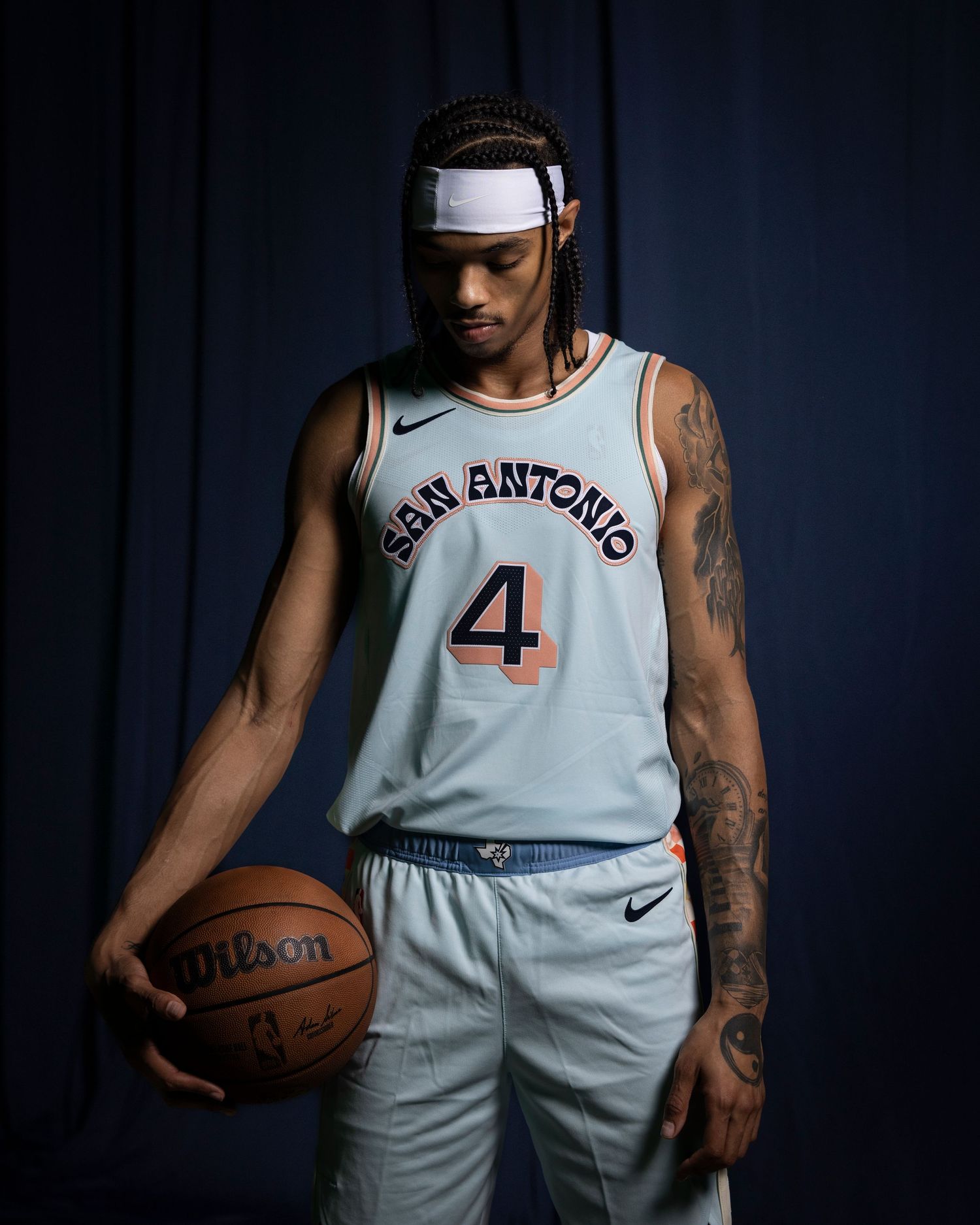 Professional basketball player poses with ball in San Antonio uniform number 4 against dramatic lighting.