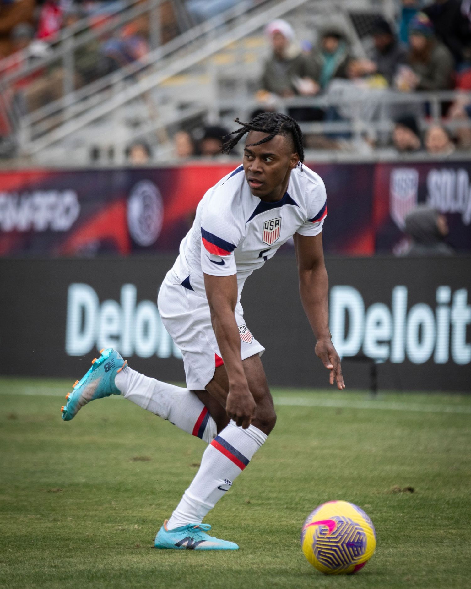 Soccer player in white uniform controls the ball on the field with Deloitte advertisement visible.
