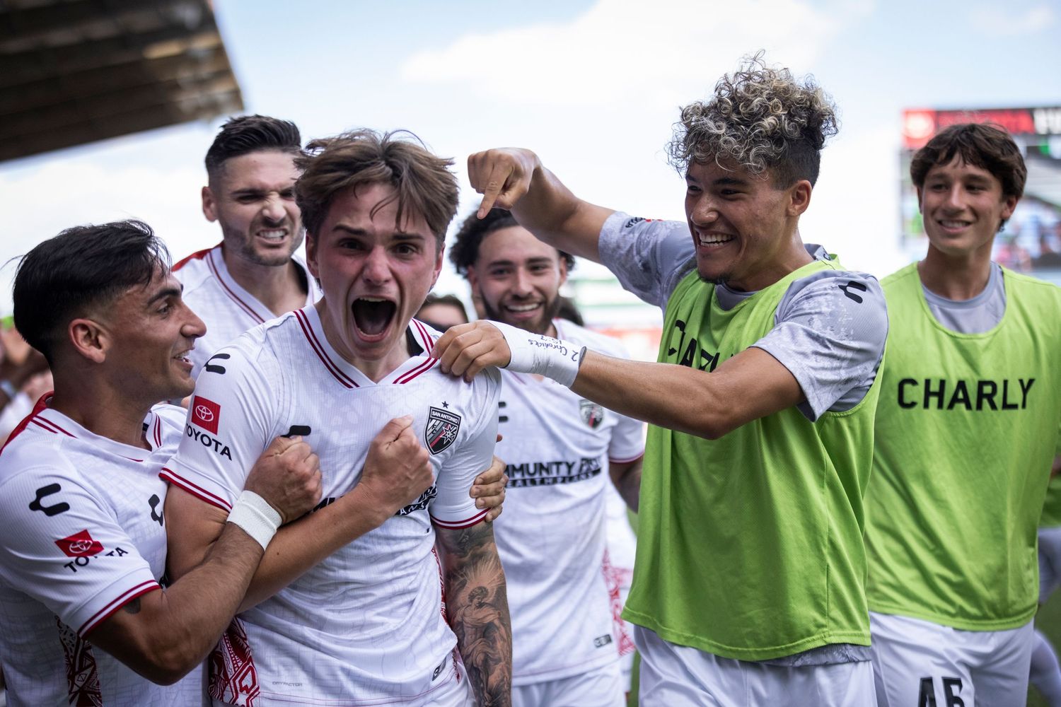San Antonio FC players celebrate joyfully in white Charly jerseys after scoring a goal during a soccer match.