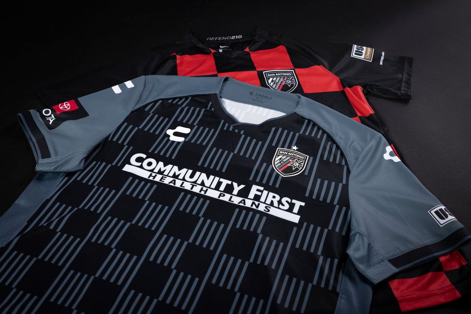 A gray SAFC (San Antonio FC) soccer jersey with Community First logo displayed on black geometric pattern background and red accents.