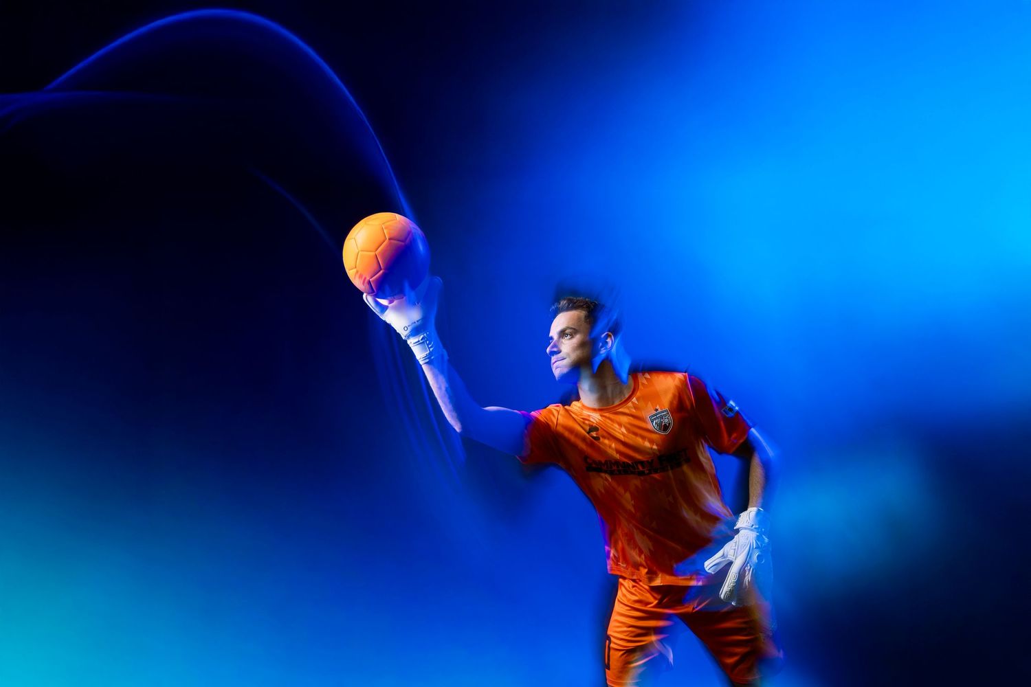 Soccer goalkeeper in orange uniform catches ball with gloved hand against vivid blue background.