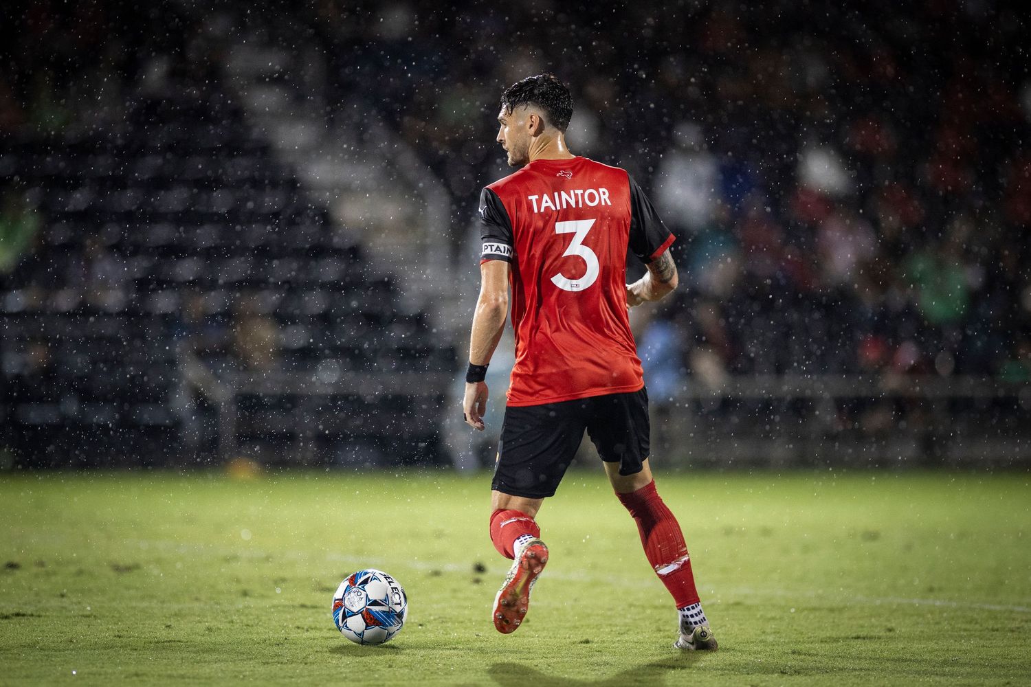 Soccer player in red jersey controls ball on field during night match.