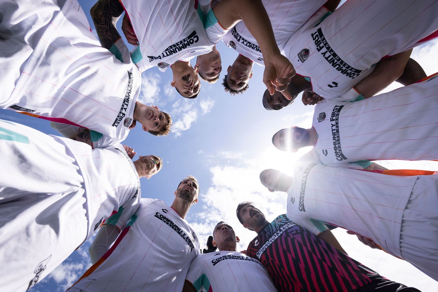 Soccer players form team huddle viewed from below with sun flare.