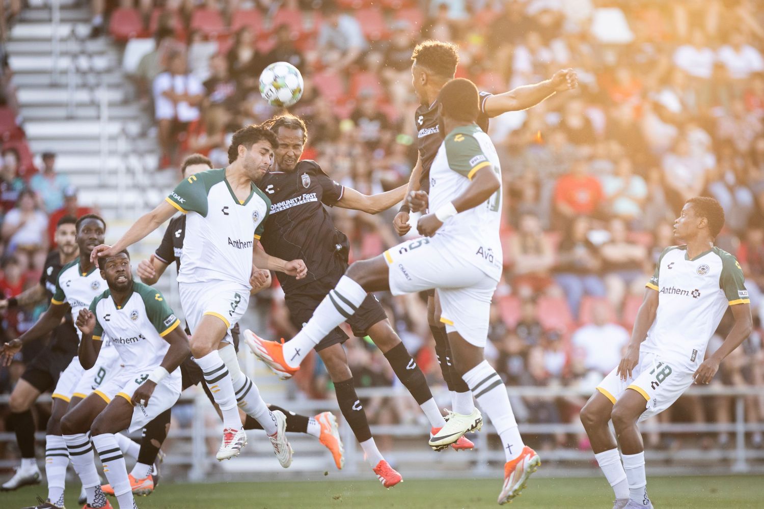 Multiple players leap high to compete for a header during a soccer match at sunset.