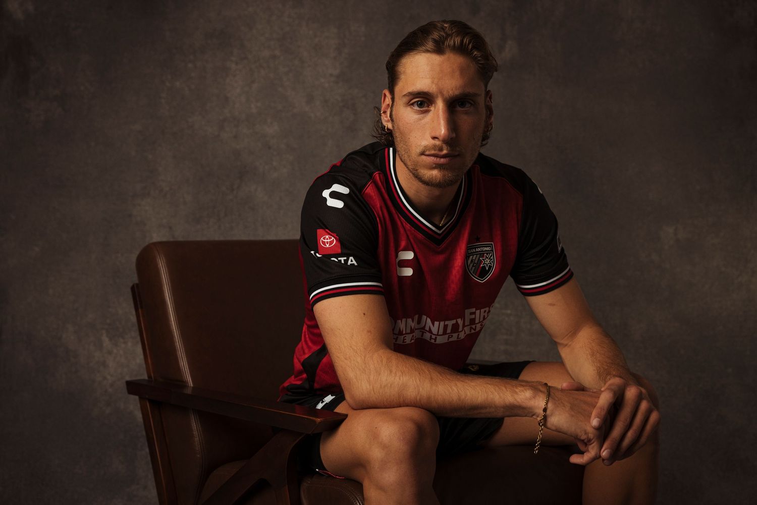 Soccer player sits confidently in a dark red and black Community First jersey during a professional photo shoot.
