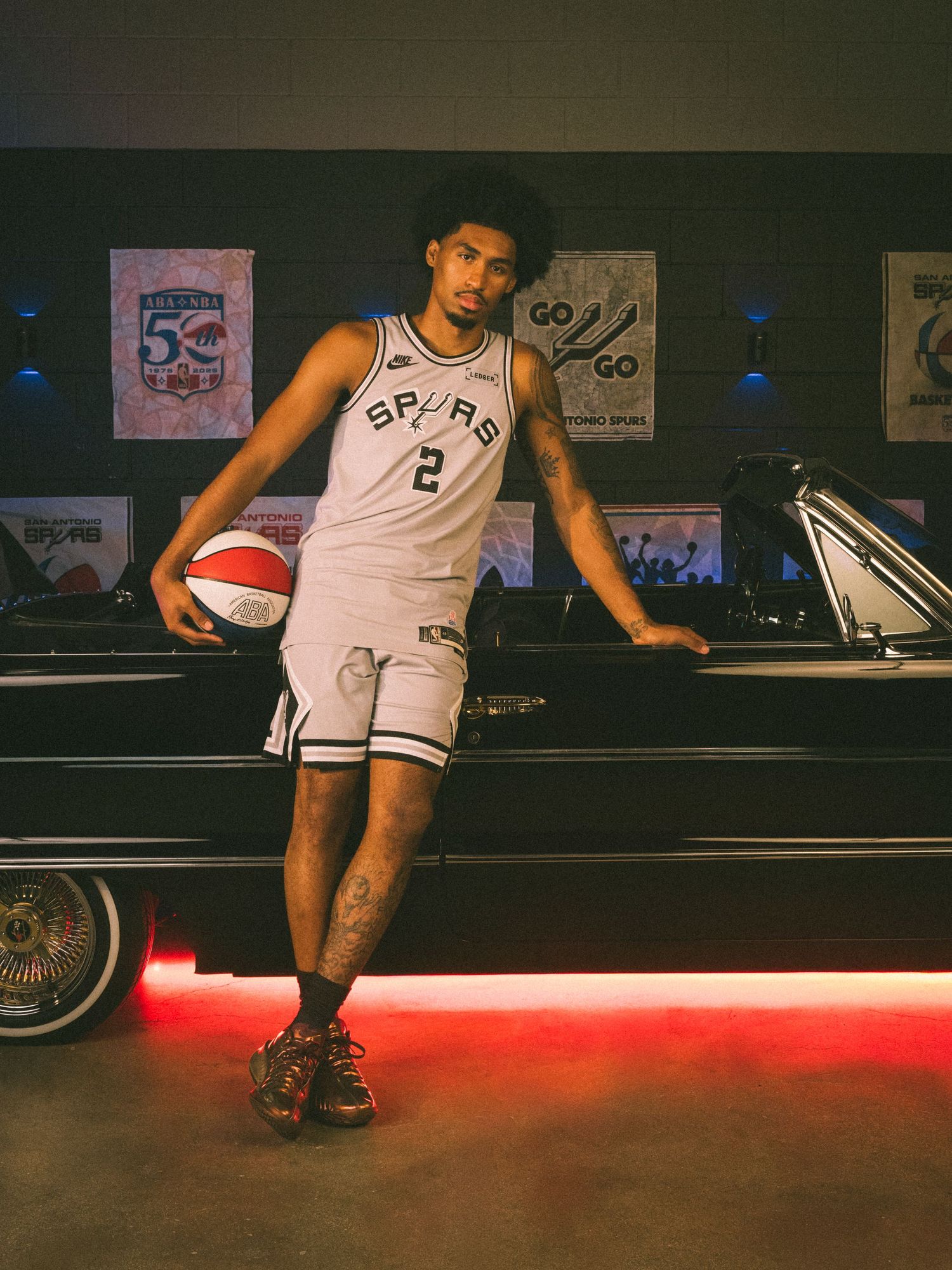San Antonio Spurs player wearing number 2 jersey poses with basketball beside a classic car in a decorated arena.