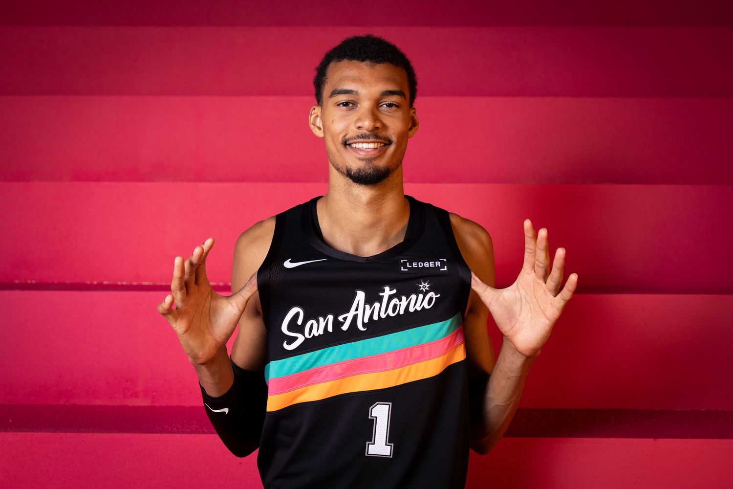 San Antonio Spurs player wearing jersey number 1 poses with hands raised in a pink-lit NBA photo session.