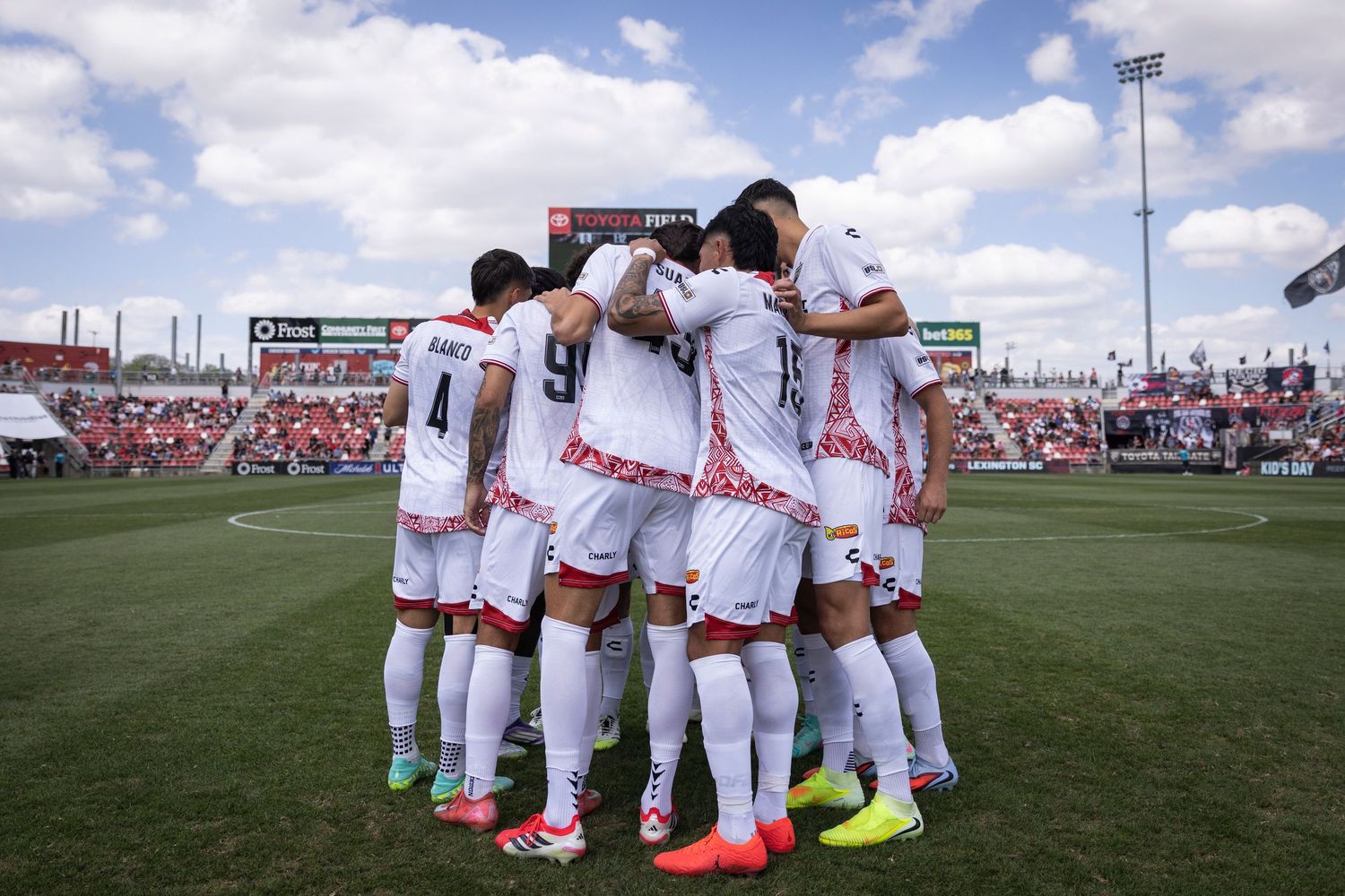 Soccer team in white uniforms forms tight huddle on field during sunny match day.