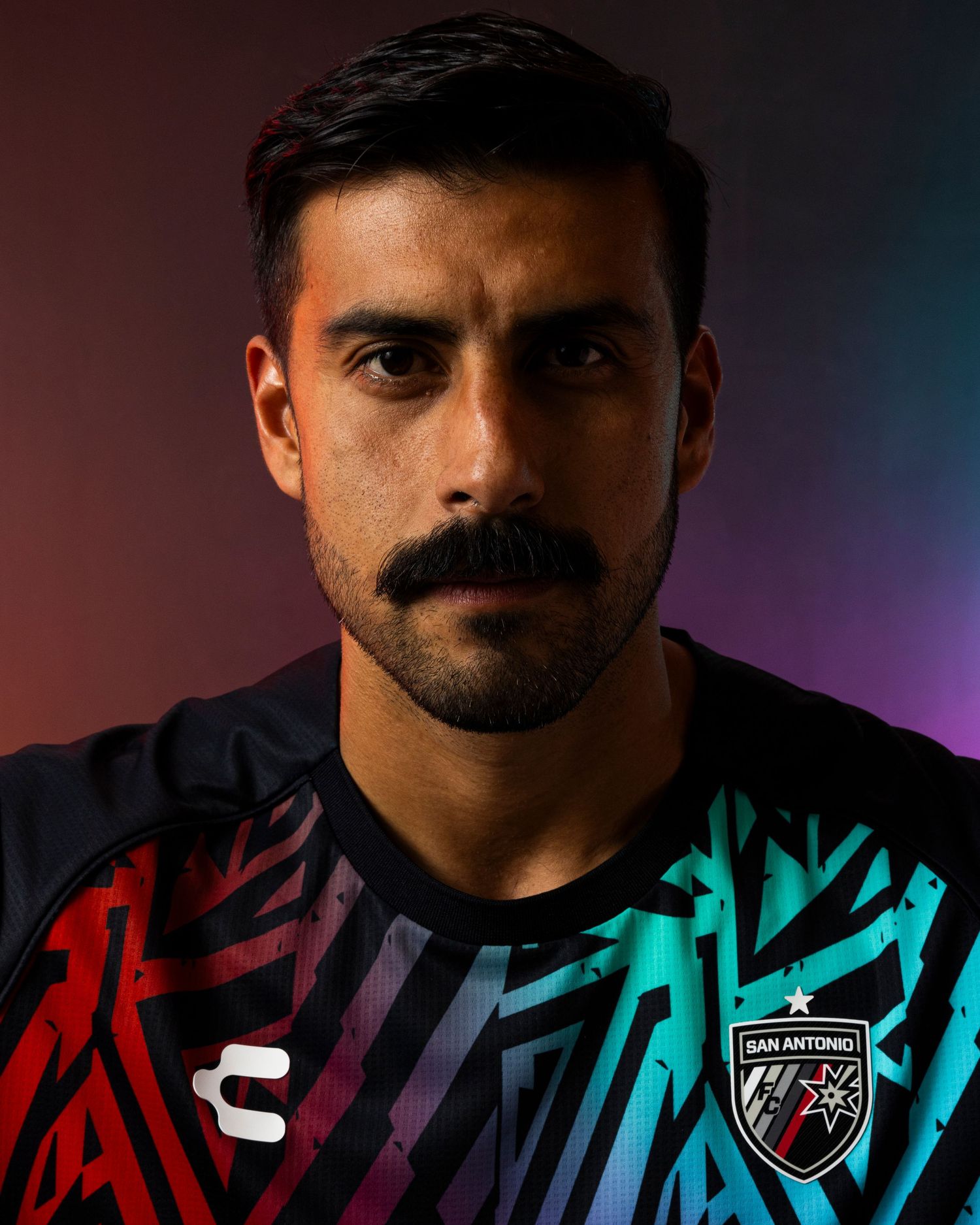 Soccer player in dramatic portrait wearing multicolored team jersey against gradient lighting.