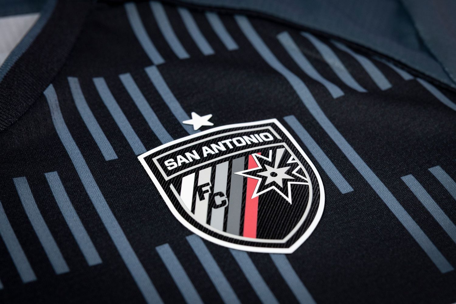 Close-up view of the San Antonio FC soccer team crest on black jersey with diagonal striped pattern. CHARLY soccer kit unveil photo