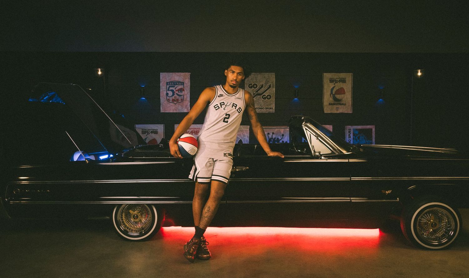 NBA player wearing Spurs jersey number 2 poses with basketball beside a classic lowrider car in a dark studio.
