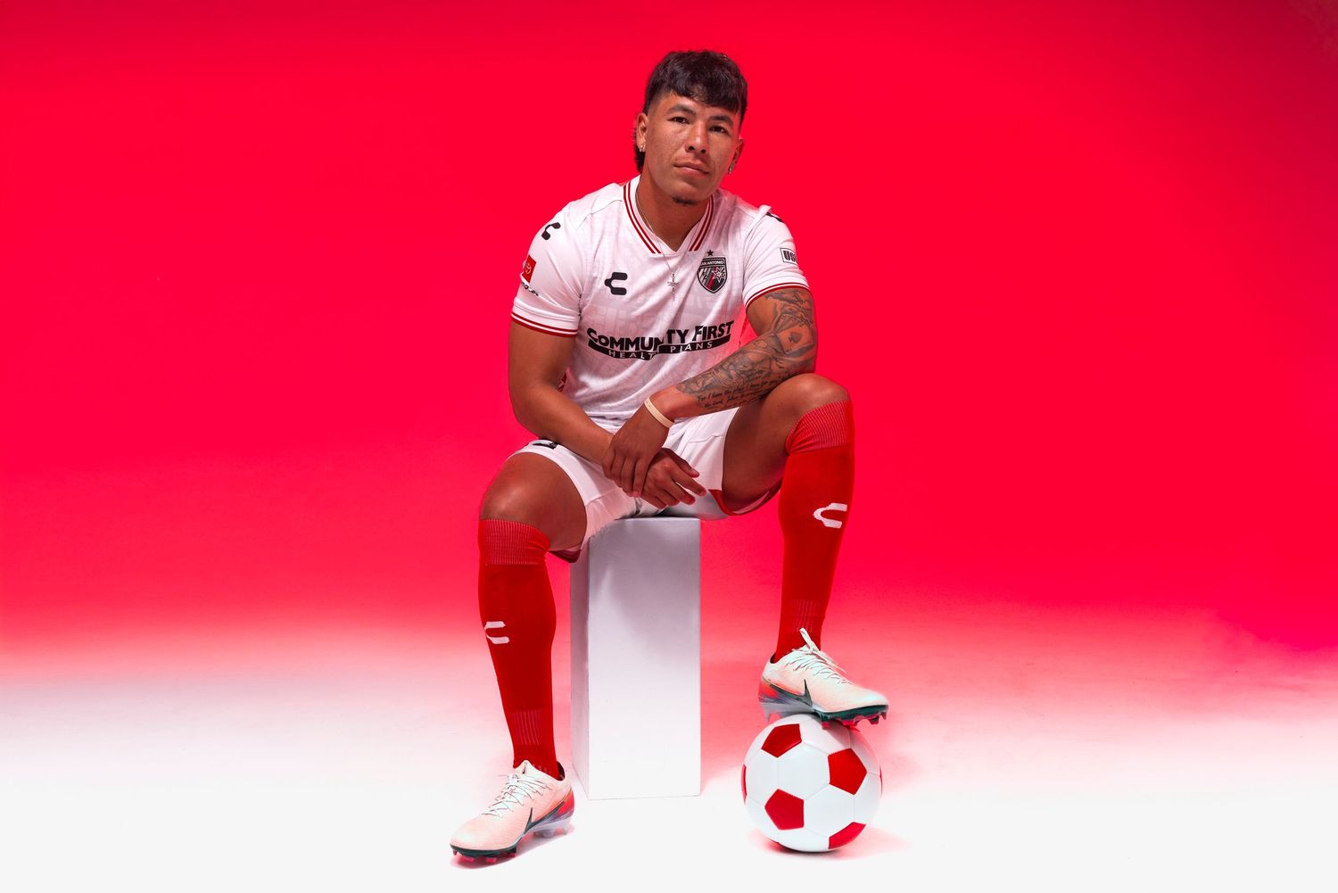 Soccer player in white and red uniform sits on cube with ball, posing against vibrant red background.