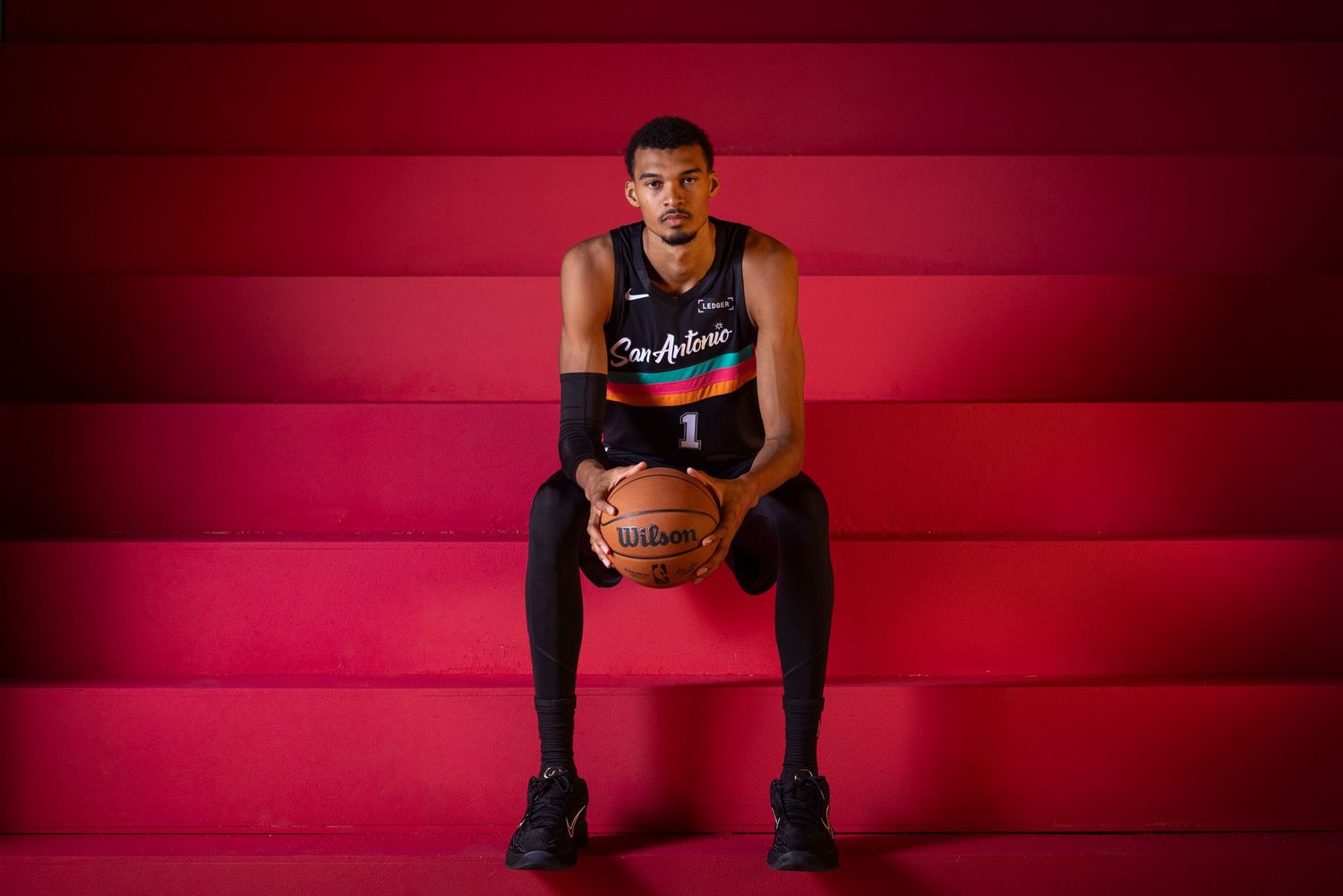 NBA player sits on red steps holding a Wilson basketball wearing a San Antonio Spurs city edition jersey.