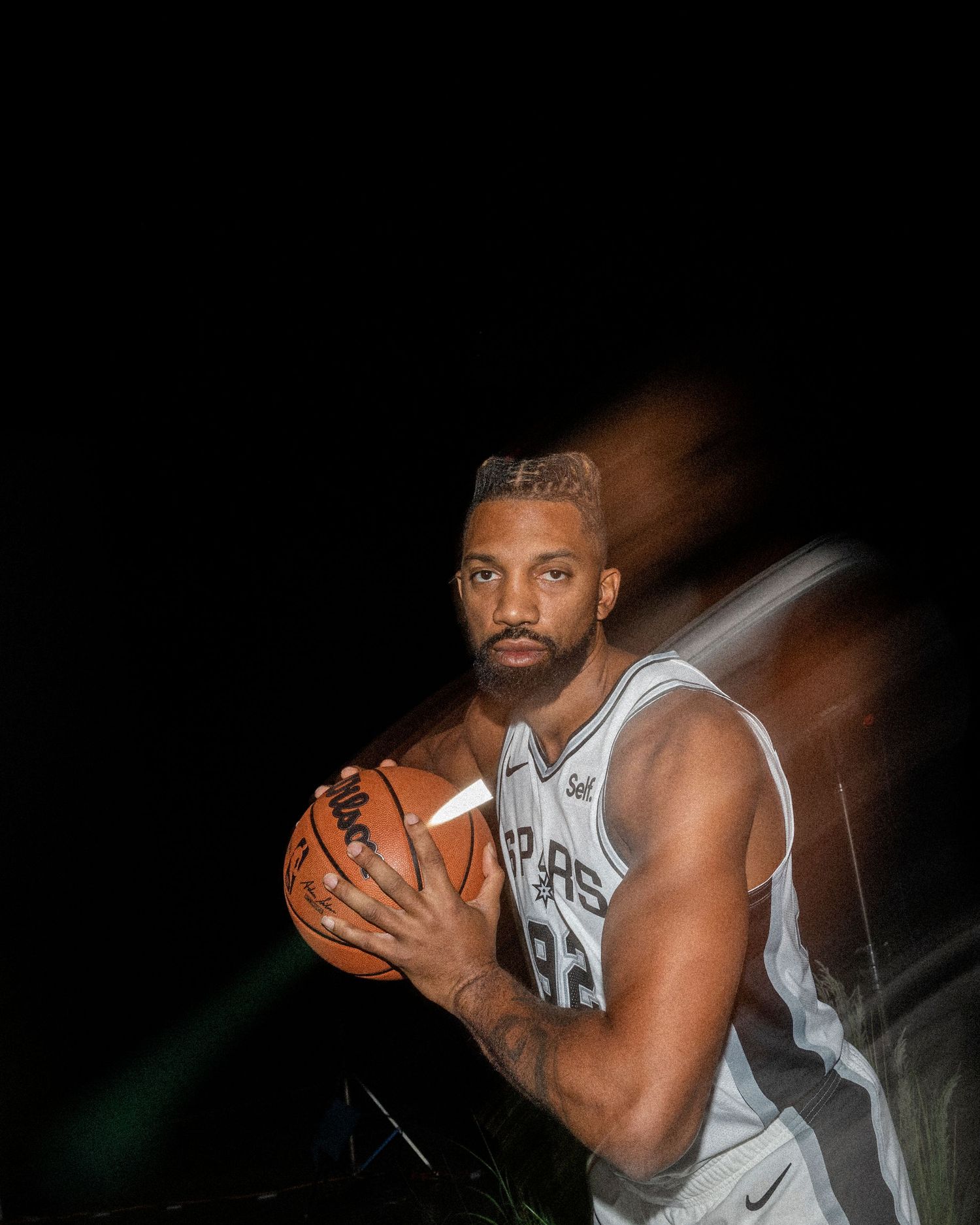 Basketball player in Spurs uniform poses with ball against dark background with motion blur effect.