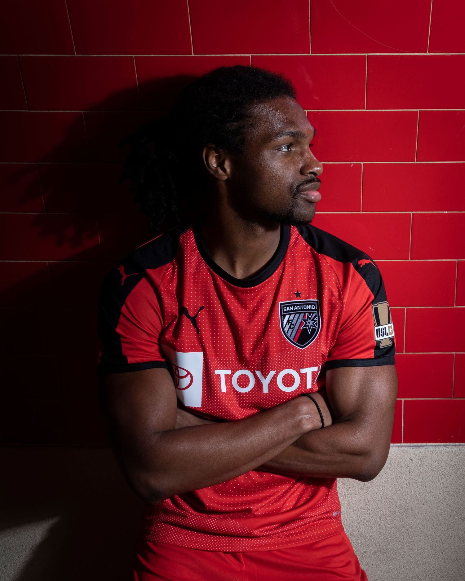 Soccer player in Toyota-sponsored red jersey poses against red tile wall.