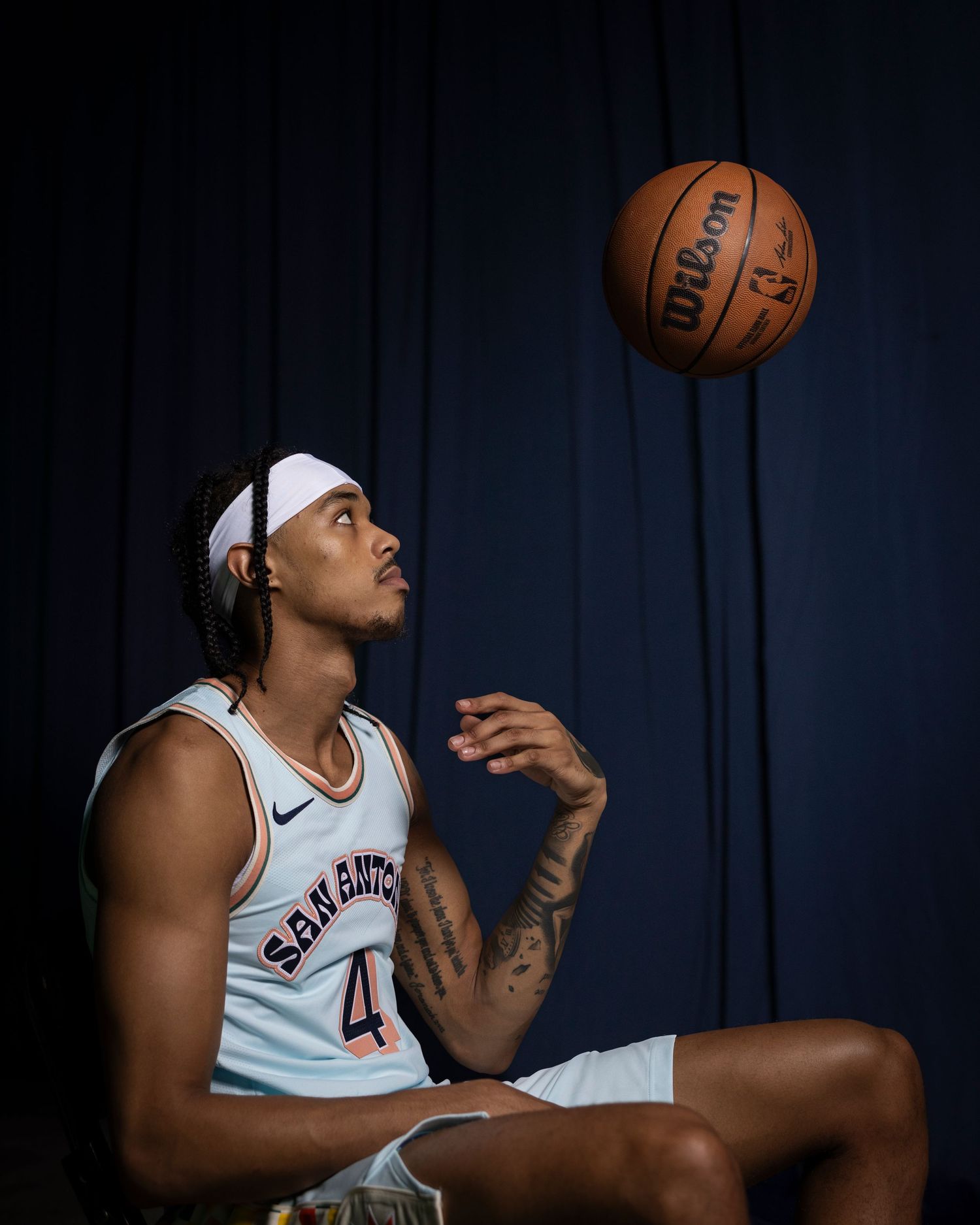 NBA player in San Antonio jersey sits contemplatively watching basketball against dark backdrop.