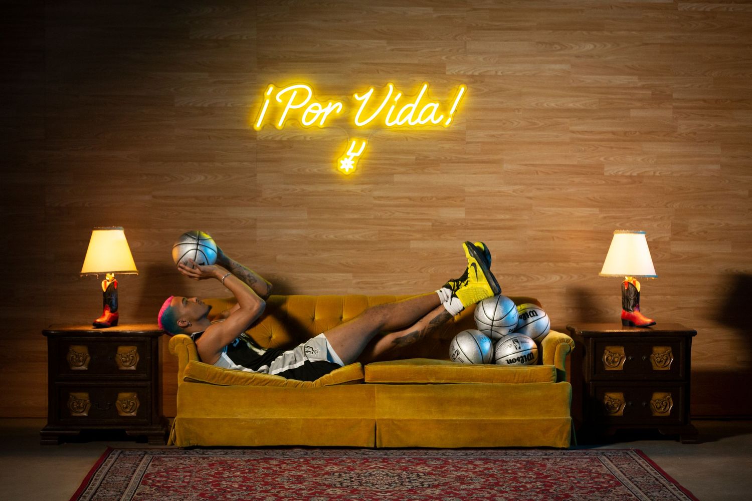 Two people lounge on a yellow couch under a neon 'Por Vida' sign with matching nightstands and lamps.
