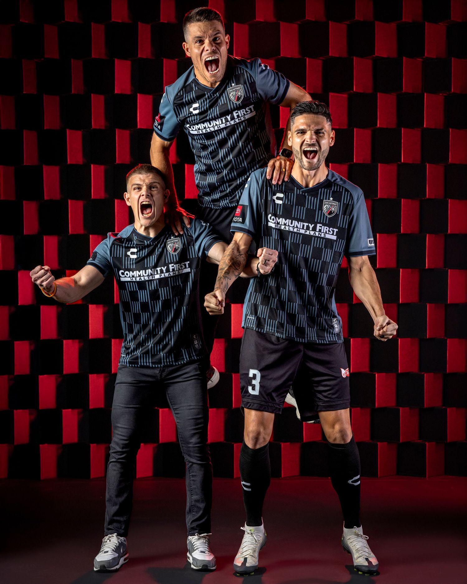 Soccer players celebrate enthusiastically in dark uniforms against a geometric red and black background pattern.