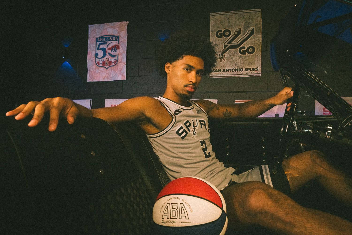 San Antonio Spurs player poses with basketball in vintage car, wearing team jersey with 50th anniversary banners in background.
