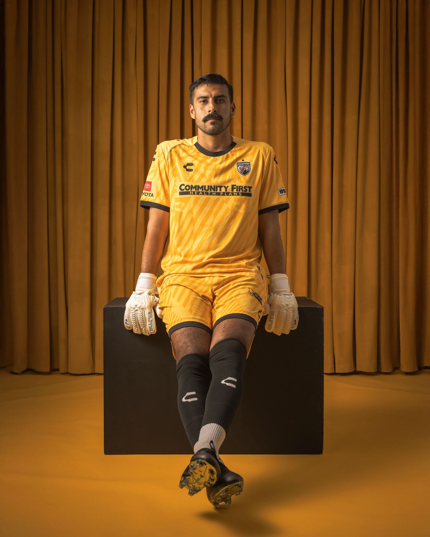 Soccer goalkeeper in yellow uniform sits on black block against brown curtain backdrop, wearing gloves and cleats.