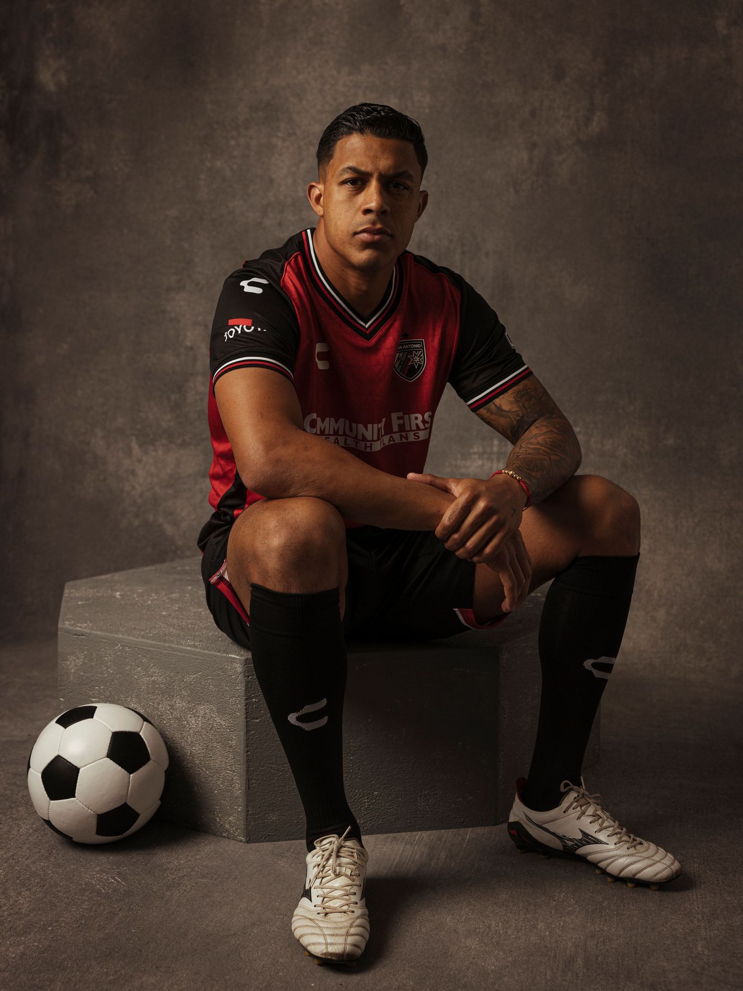 Soccer player in red and black uniform sits on a cube beside a soccer ball in a studio portrait.