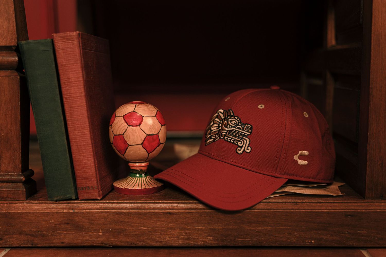 Burgundy Charly baseball cap beside a decorative soccer ball on a wooden shelf with books.