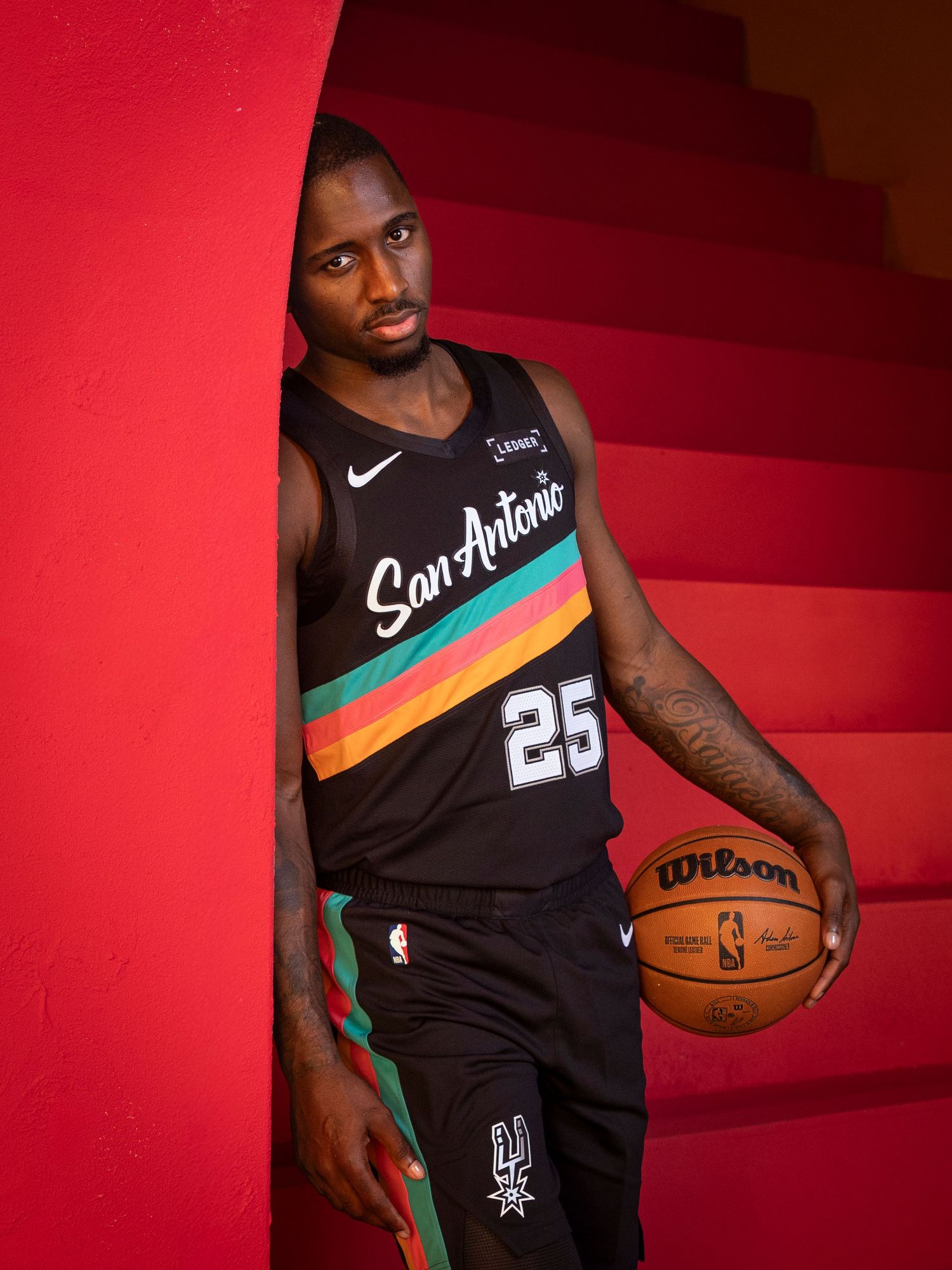 San Antonio Spurs player #25 holds a Wilson basketball on a red staircase in black City Edition jersey.