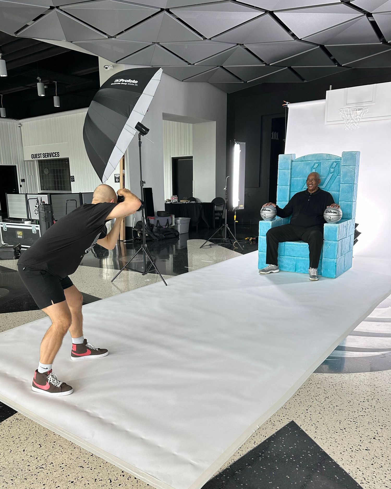 Behind-the-scenes photo shoot setup in a studio with lighting equipment, white backdrop and turquoise chair.