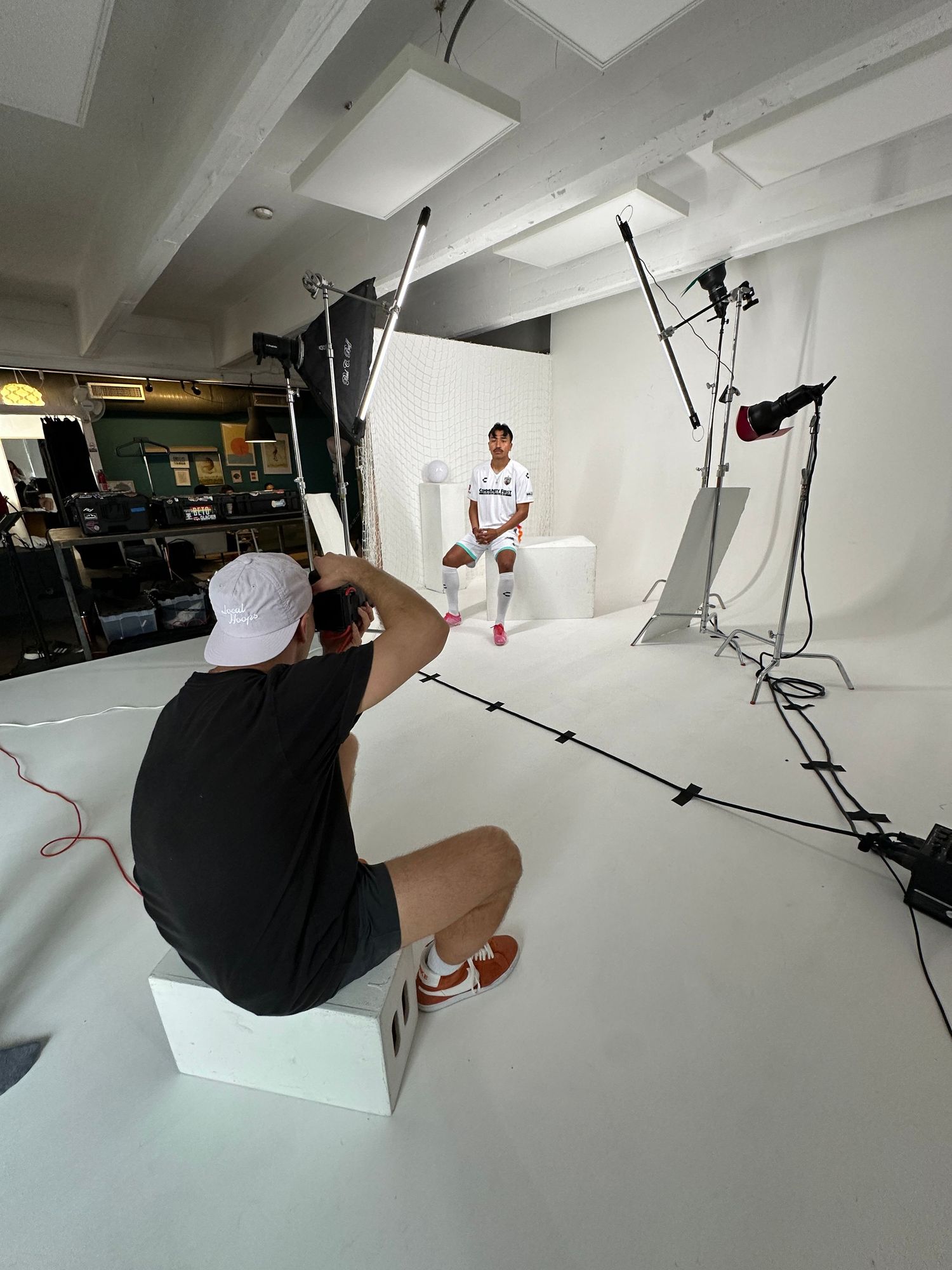 Behind-the-scenes photo of a professional photoshoot in a white studio with lighting equipment and photographer in action.