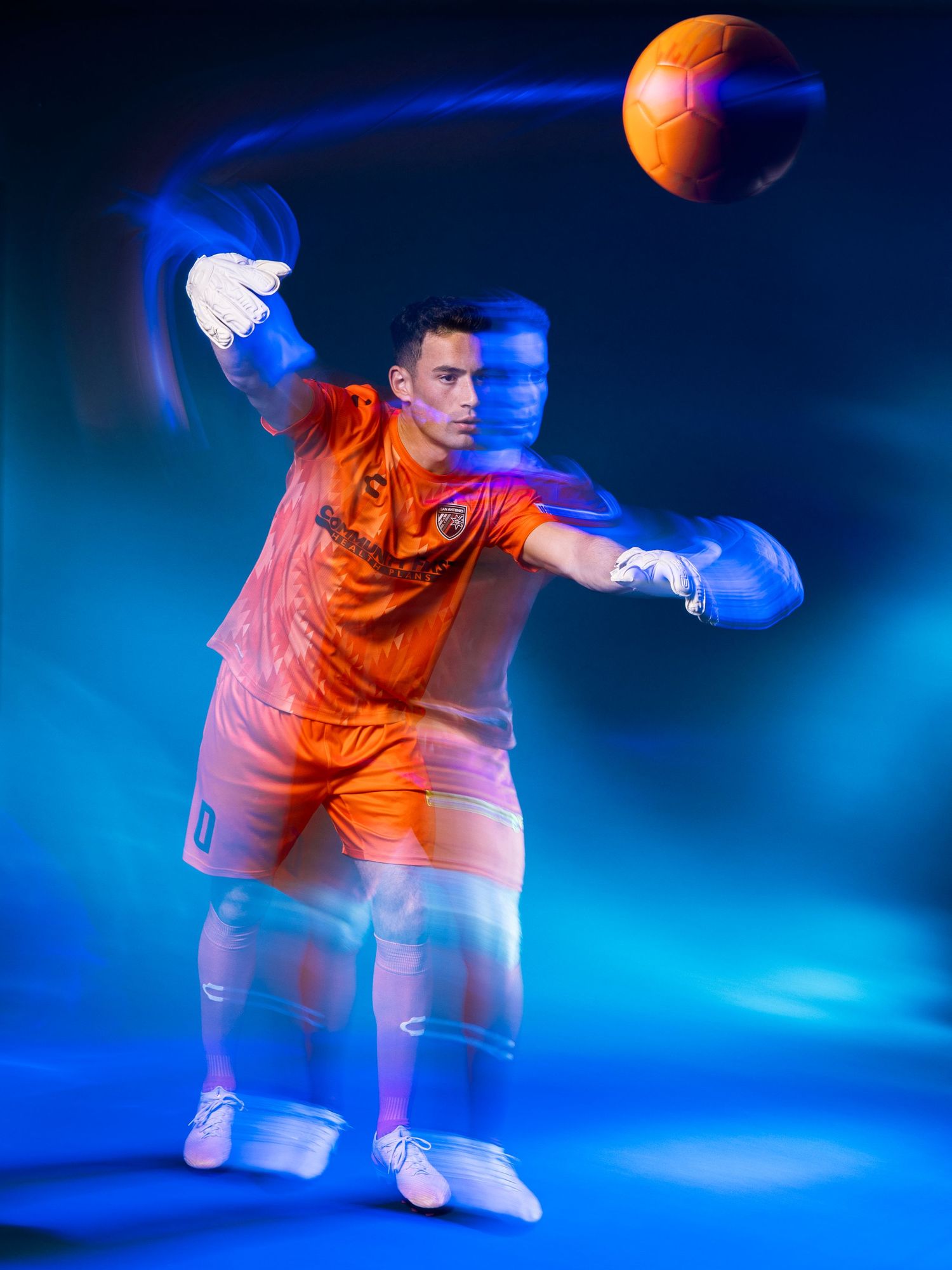 Soccer goalkeeper in orange kit makes dynamic save with motion blur effect against blue background.