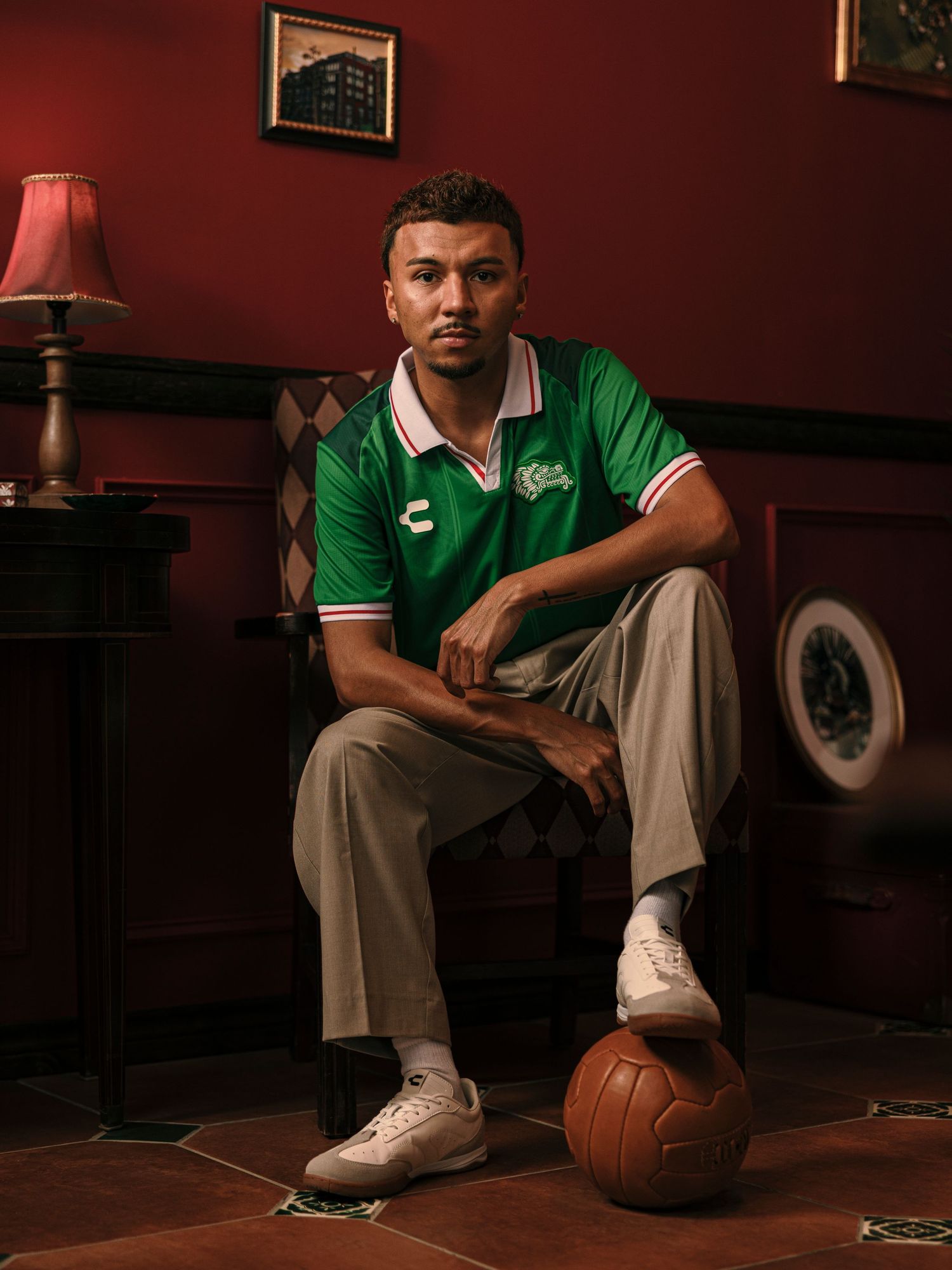 Man in green Chchupa soccer jersey sits confidently with vintage football at his feet in a moody red room.