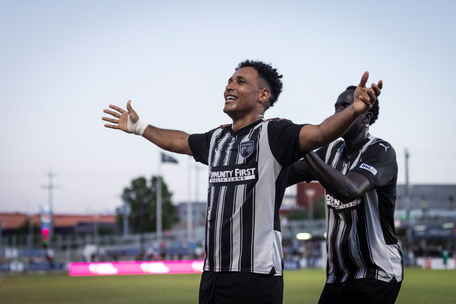 A soccer player celebrates with arms outstretched while wearing a black and white striped jersey.