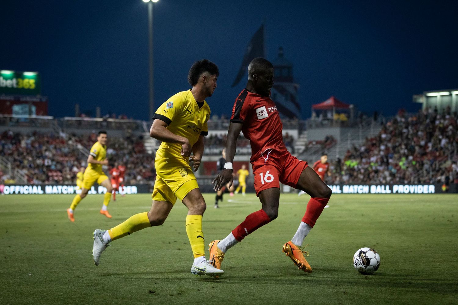 Soccer players from opposing teams battle for possession during nighttime match under stadium lights.