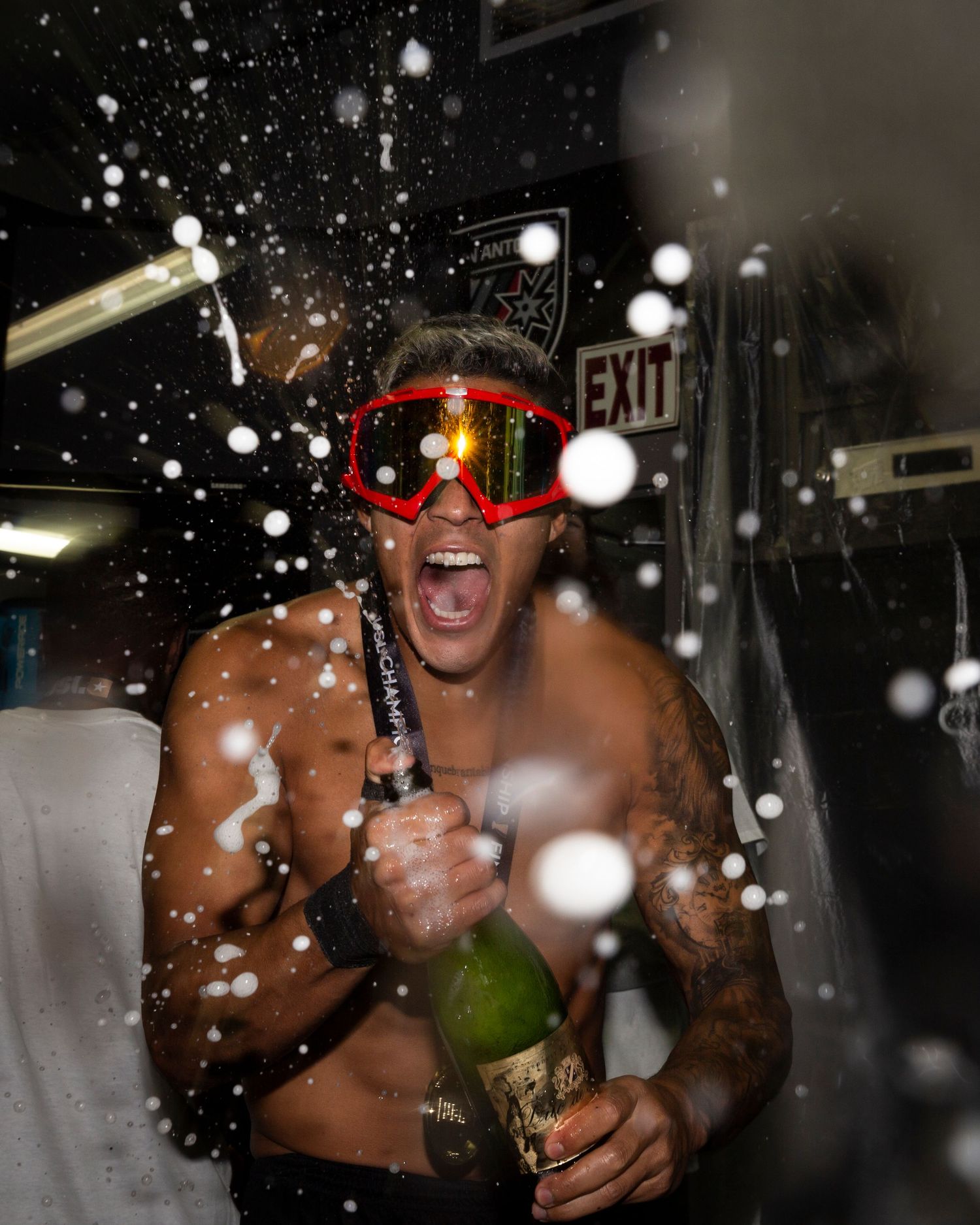 Athlete celebrates with champagne spray while wearing red goggles in dark setting.