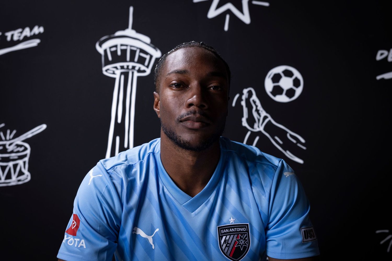 Soccer player in light blue jersey poses against wall with sports-themed illustrations.