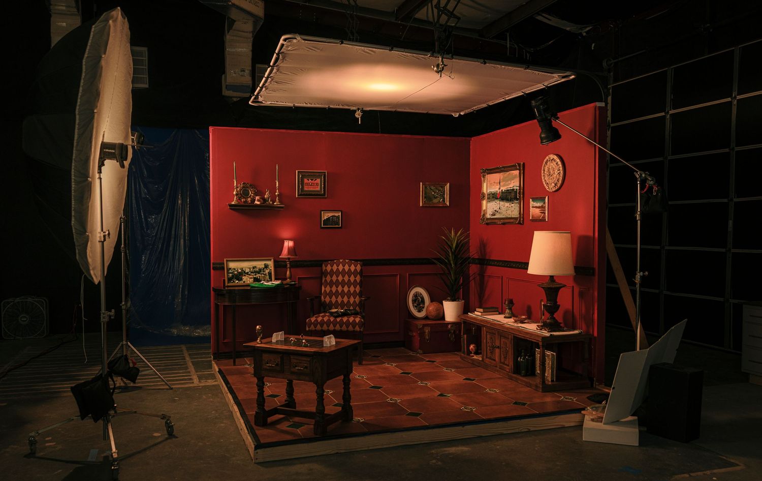 Dark studio lighting setup with red walls and professional photography equipment arranged around a vintage-style interior scene.