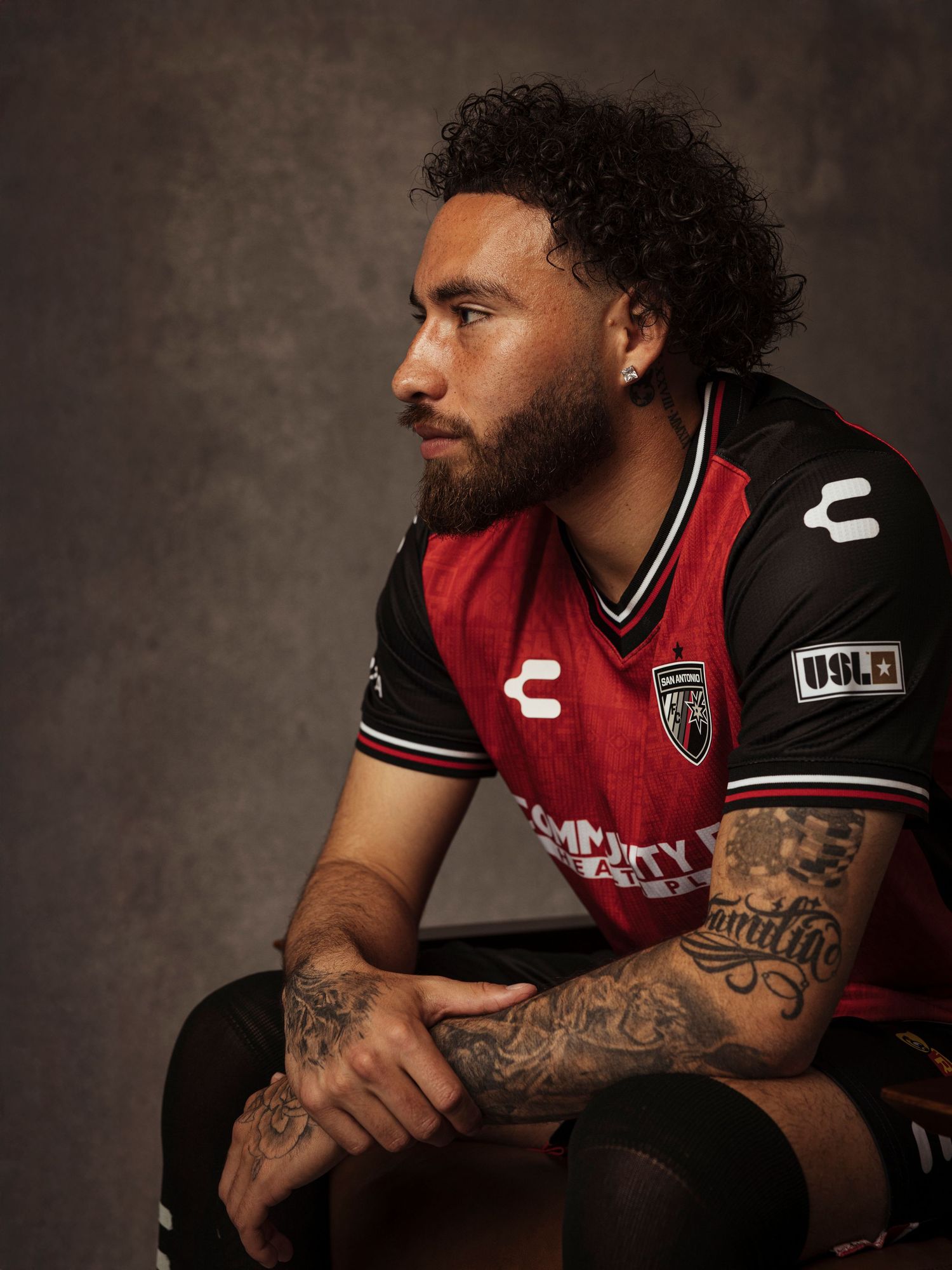 Tattooed soccer player in red and black Charly USL jersey sits pensively in a studio portrait.