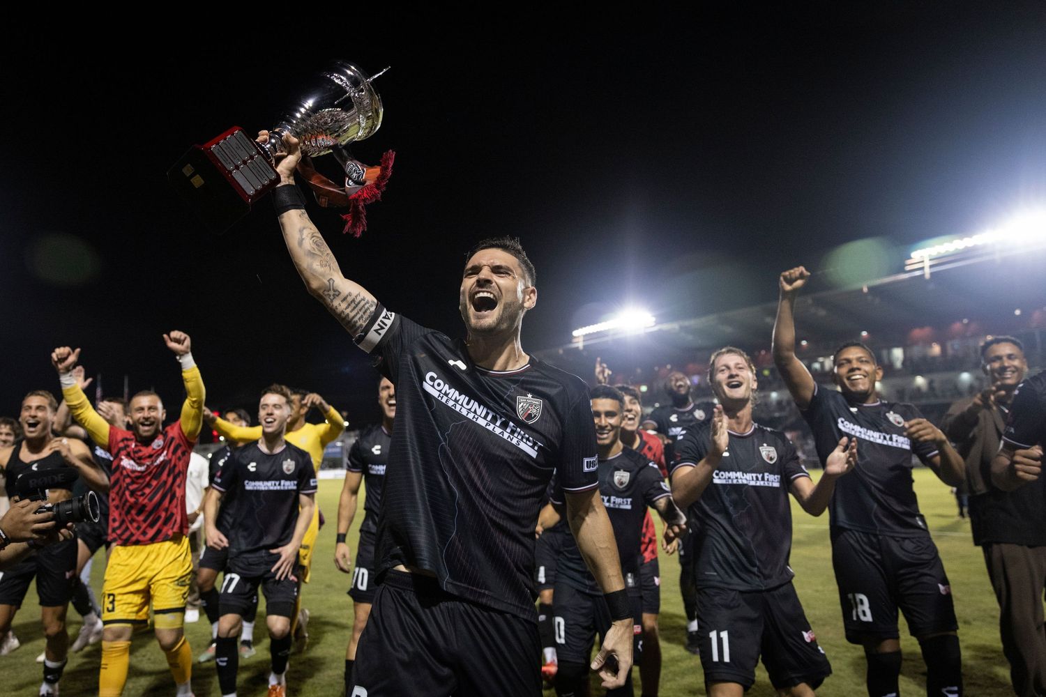 Soccer team celebrating championship victory, player lifting trophy triumphantly under stadium lights.