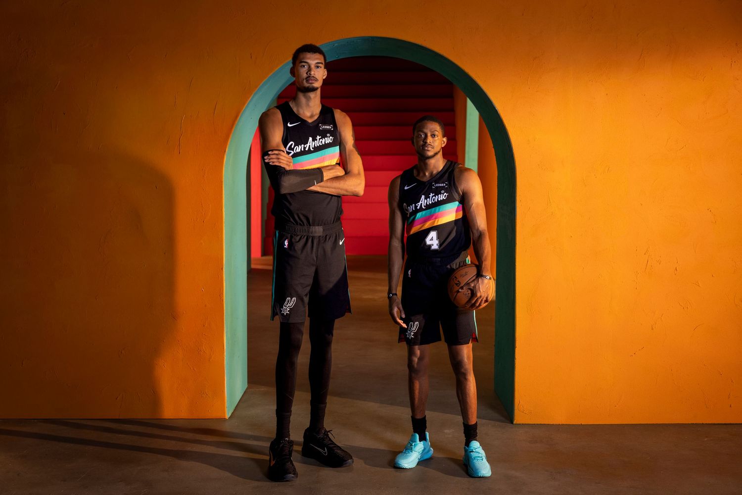 Two basketball players in Spurs uniforms pose dramatically against a vibrant orange and teal backdrop.