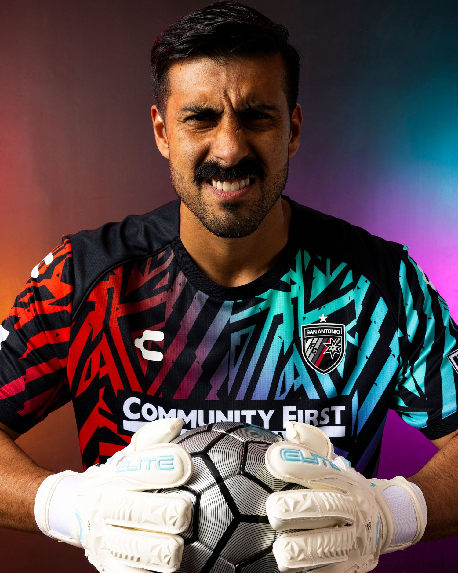 Goalkeeper in colorful striped jersey poses with white gloves against gradient background.