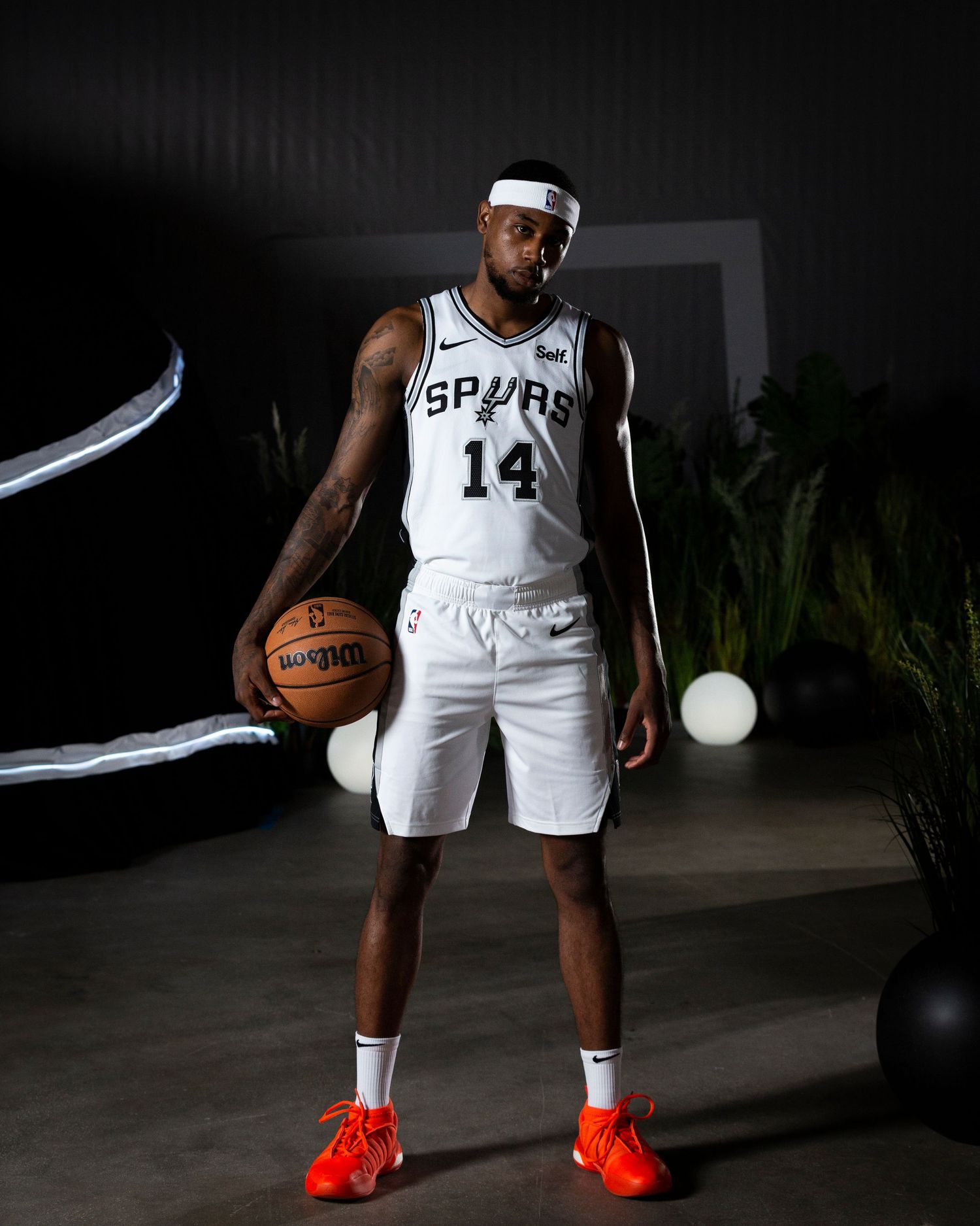 Basketball player in Spurs number 14 jersey stands with ball in studio setting with decorative plants.