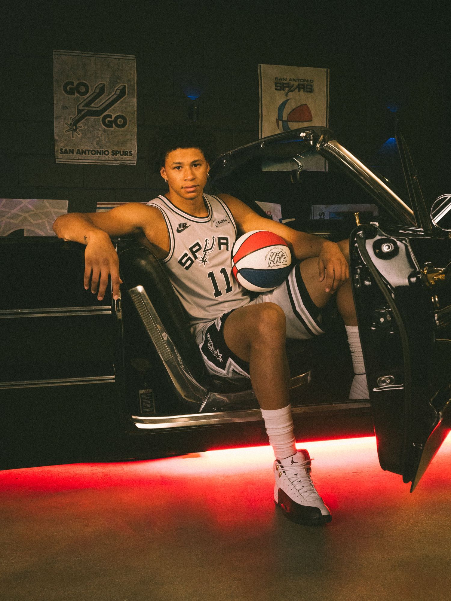 San Antonio Spurs player #11 poses with a basketball beside a car with red underglow lighting.