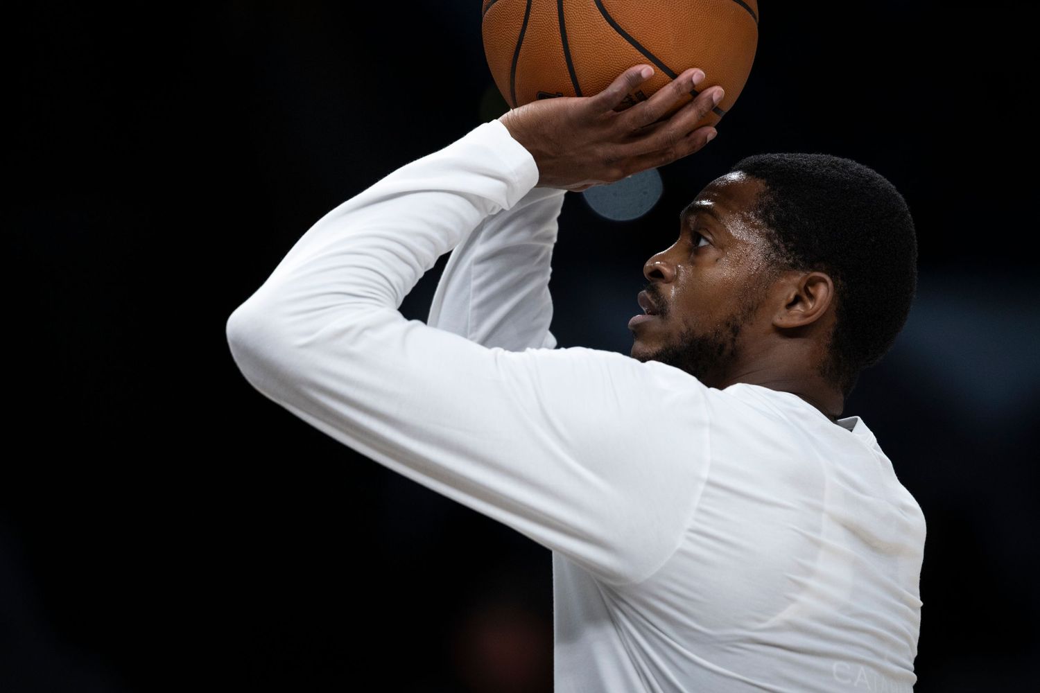 Basketball player in a white long-sleeve warm-up shirt raises a basketball overhead while taking a shot in a dark arena.