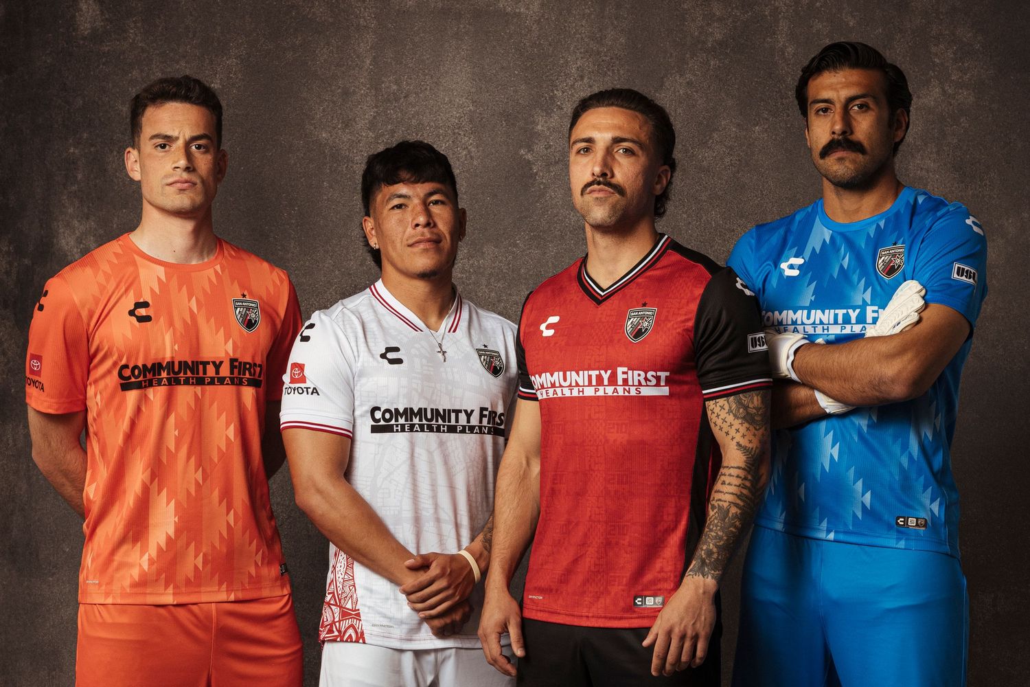 Four soccer players pose in colorful jerseys including orange, white, red, and blue Community First sponsored kits.