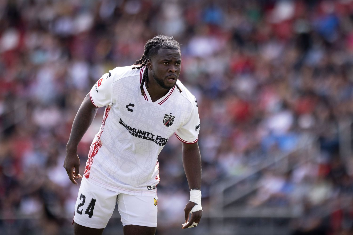 Soccer player wearing number 24 white jersey stands on field during a daytime match with crowd in background.