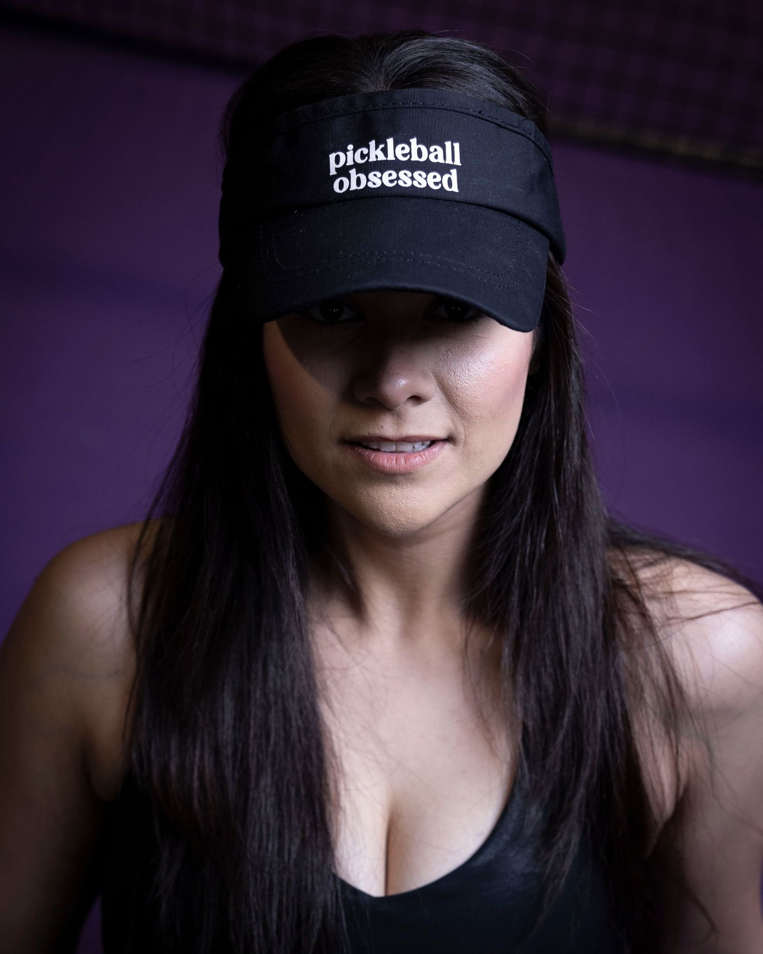 Close-up portrait of person wearing black 'Pickleball Obsessed' cap against purple background.